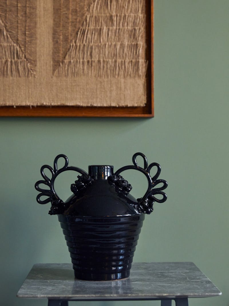 Black glossy glazed terracotta vase by Italian designer Valentina Cameranesi sitting on marble table top with work by Sophie Rowley in the back hanging on a green wall.