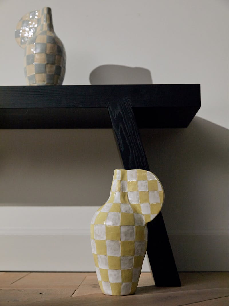 Two checkered ceramic objects by Maria Lenskjold, one is white and yellow and the other is blue and beige. They sit on a black wooden bench and on a wooden floor.