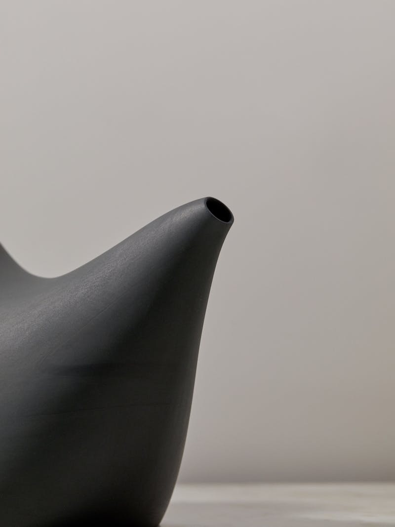 detail image of dark grey charcoal coloured porcelain vessel by Aldo Bakker. The spout of the vessel is tapered and resembles the small side of a pipe. 