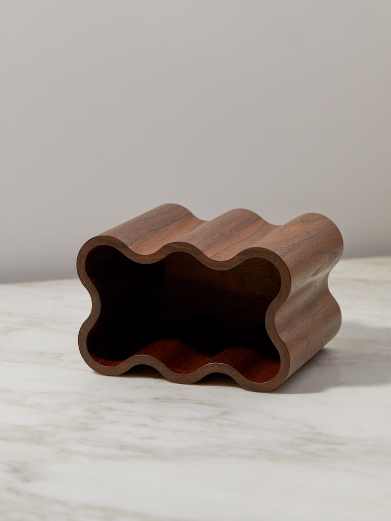 sculptural walnut wooden container by Jonas Lutz, Finnish-born designer based in Rotterdam. The box has a wavy edge and a smooth surface and is shown laying on its side on top of a marble table top.