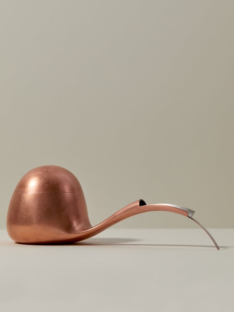 copper and silver curved soy pouring vessel with a long sculptural neck and rounded dome like body. The pourer is shown on a white surface and is made by Dutch designer Aldo Bakker. 