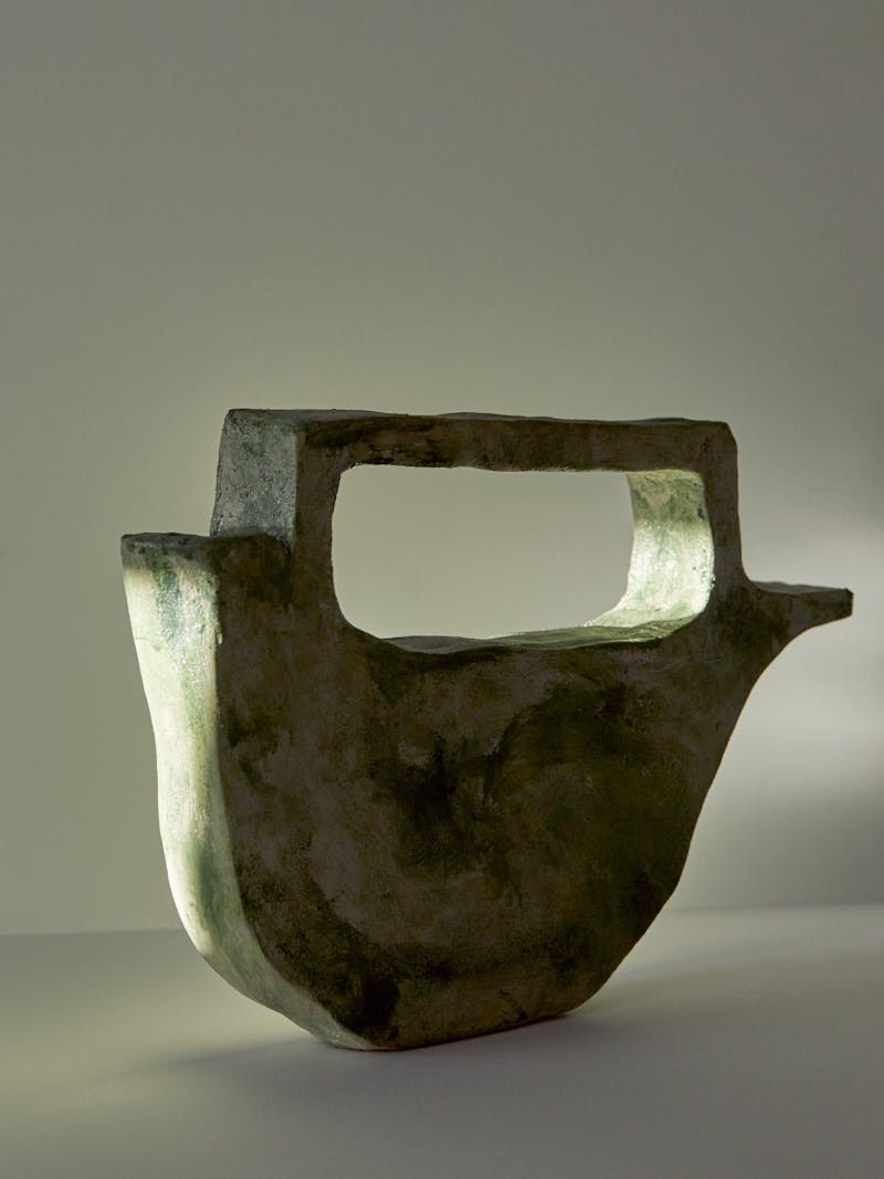 high contrast and shadowy image of a green flat ceramic vessel with a wide cut out handle by Willem van Hooff. The base of the sculptural vessel is curved and it has a spout.