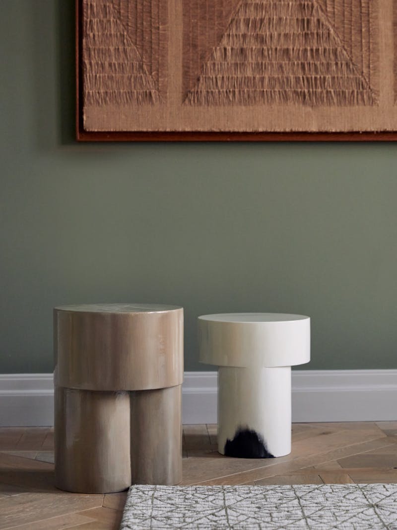 pair of collectible horn side tables from lacquer by destroyers/builders, one is in taupe and the other is in white with black detailing. The stools sit on a wooden floor with a wall work by Sophie Rowley behind them. 