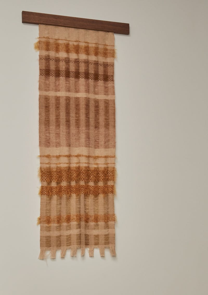 A hand woven wall hanging textile by Wovenform with colours of orange, pink, brown and cream. The fiber piece is hung on the wall with a piece of dark wood. 