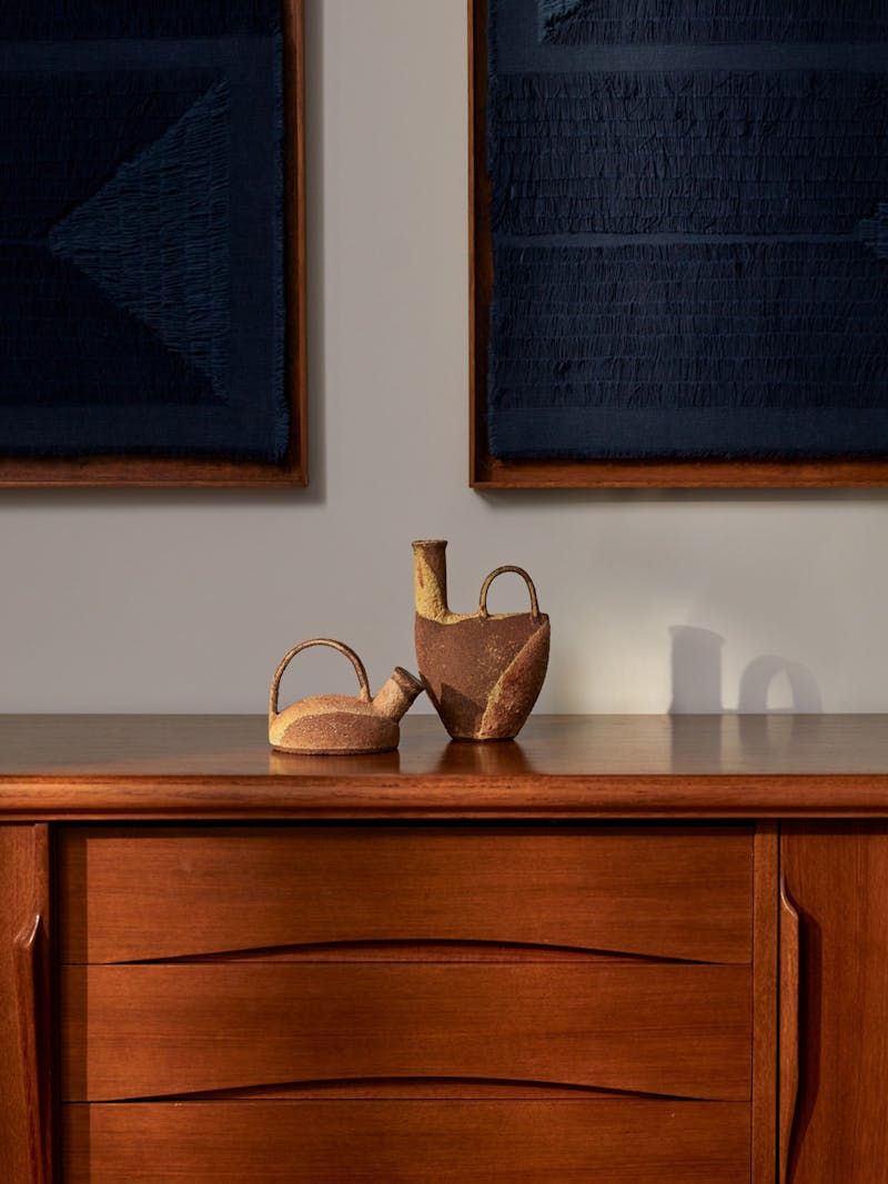 Installation image of a set of stoneware ceramic vessels by Catherine Dix with caramel, brown, cream and rust red colours, each vessel has an earthy texture includes a handle and pouring spout and sits on top of a wooden cabinet with blue textile wall works by Sophie Rowley in the background.