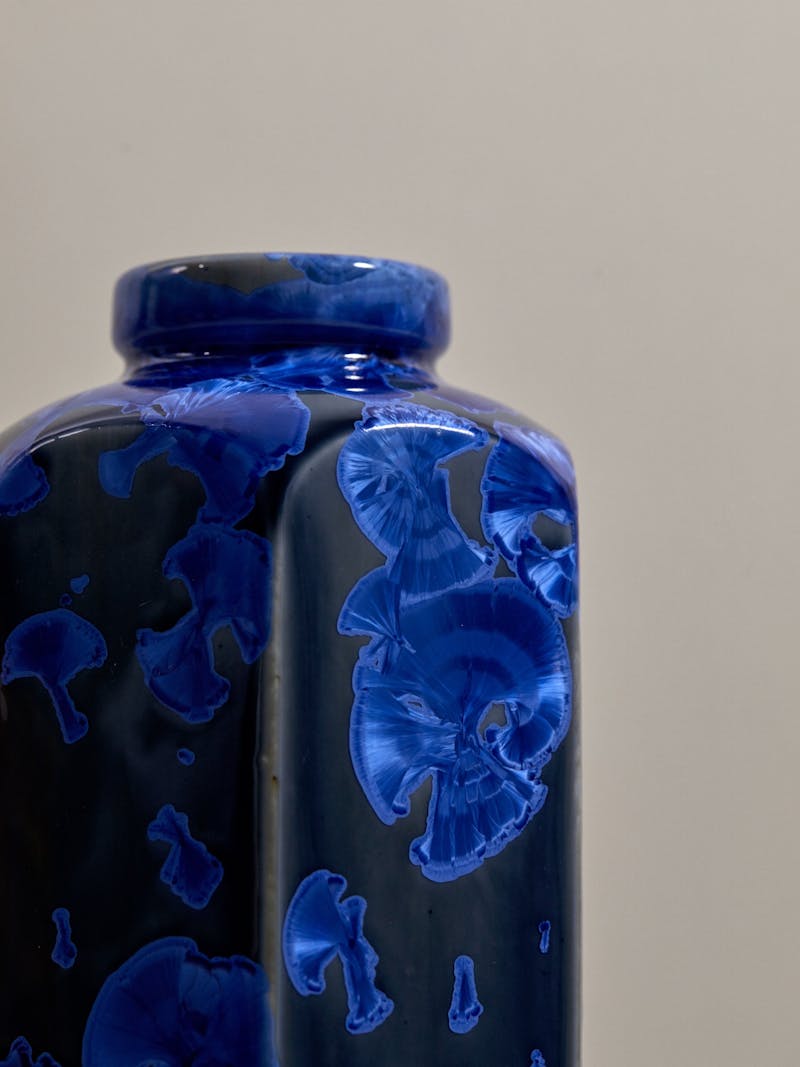 A detail image of a handmade blue hexagonal vase. The porcelain vessel is by Milan Pekar and has a dark blue background with a royal blue crystalline iridescent glaze over it. 