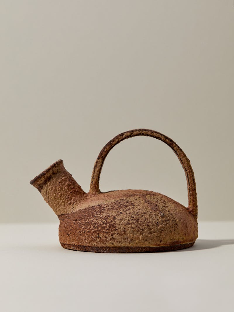 profile image of a stoneware ceramic vessel by Catherine Dix with handle and pouring spout. the clay is fired in a combination of caramel, brown, cream and rust red colours. the sculptural vessel sits on a white table top.