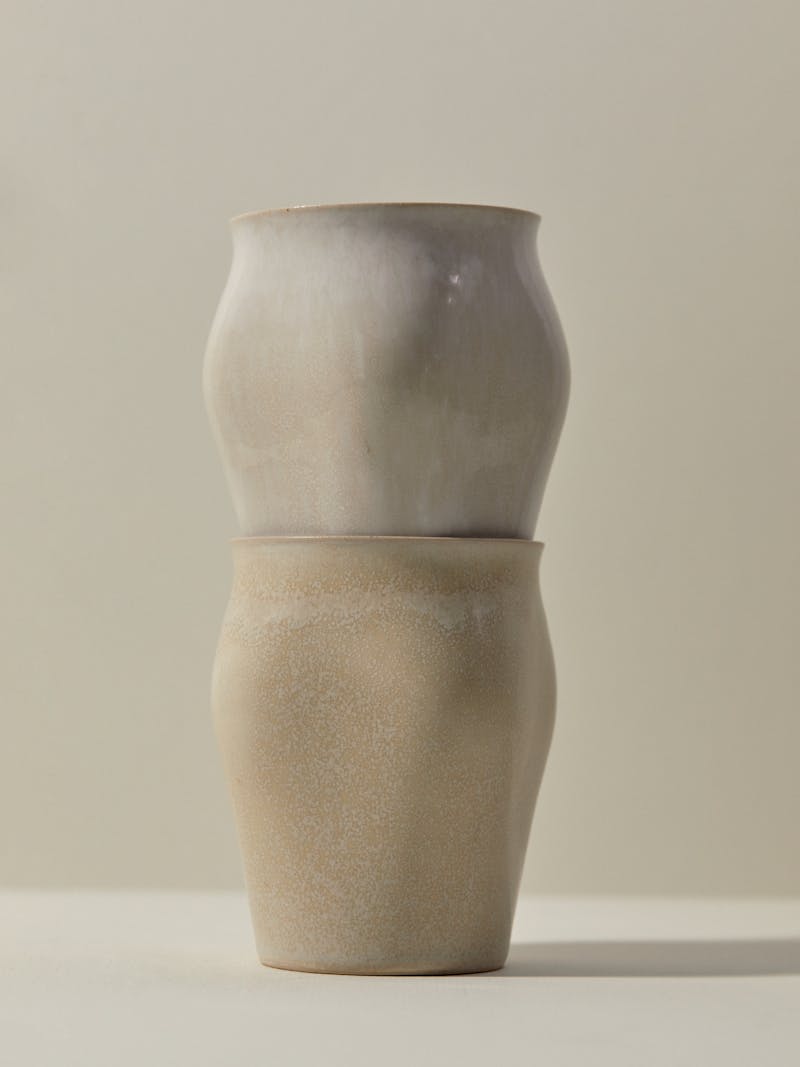 Two unique handmade cream coloured ceramic curved cups are shown stacked on top of each other from Russian designer Maria Tyakina. The vessel slip cast and glazed and sits on a white table top.