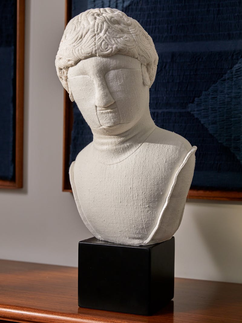 off white sculptural textile bust by Spanish designer Sergio Roger sitting on a wooden surface with a dark blue textile work by Sophie Rowley in the background. 