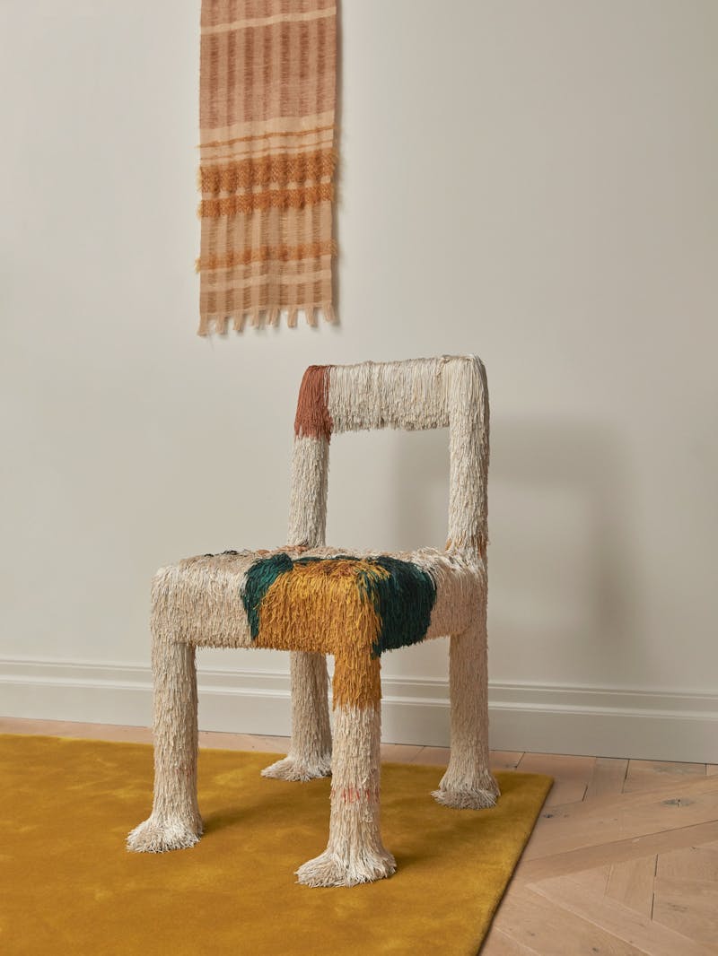a fringed chair by Buro Belen with wool cut pieces. The hair of the chair are in beige, ochre, blue and orange. The chair sits in front of a fiber piece from Wovenform and on top of a gold ochre coloured rug.