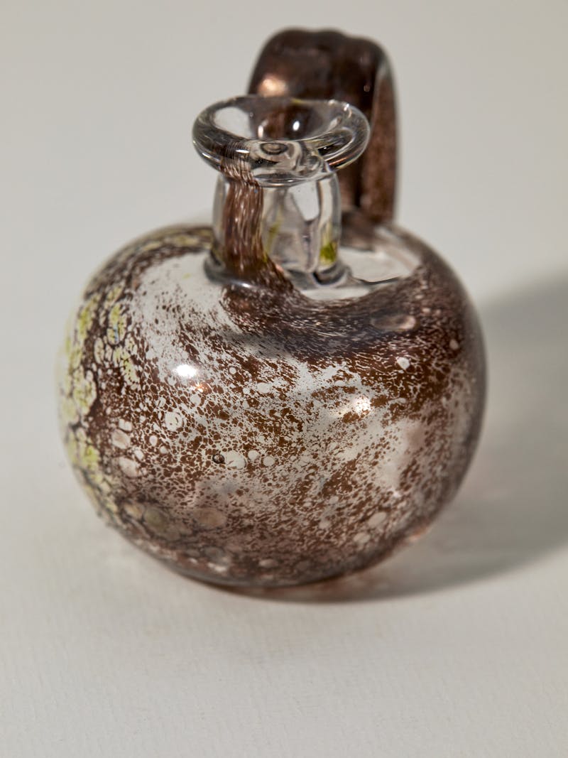 A glass vessel handblown by artist Pauline Bonnet, the glass is clear with swirls of brown and green speckles inside of it. There is a single handle, a wide round base and a spout with a lip. 