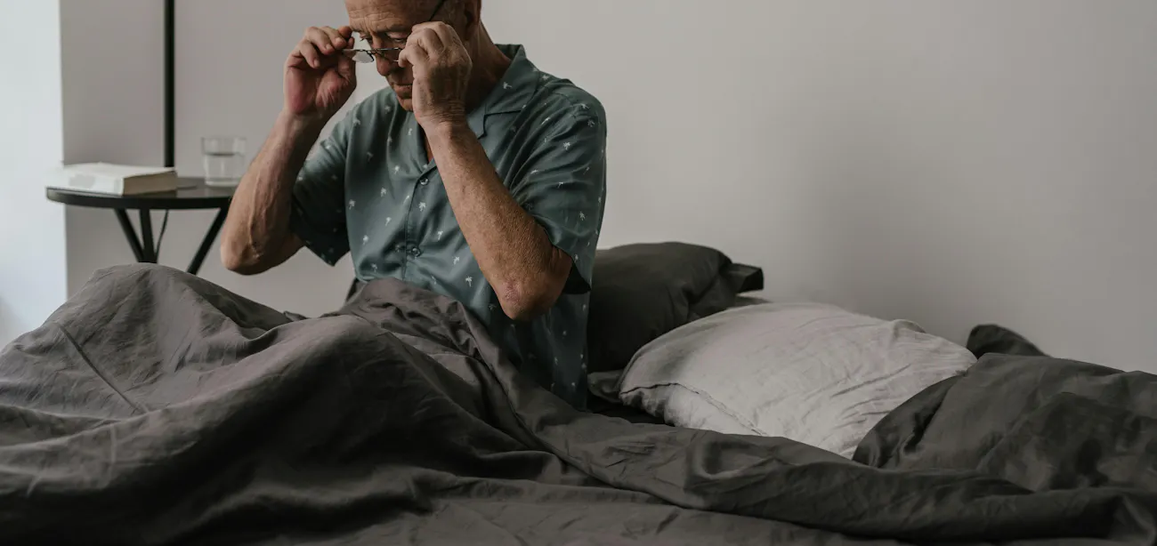 A man sitting on the edge of a bed to wipe sweat from his forehead
