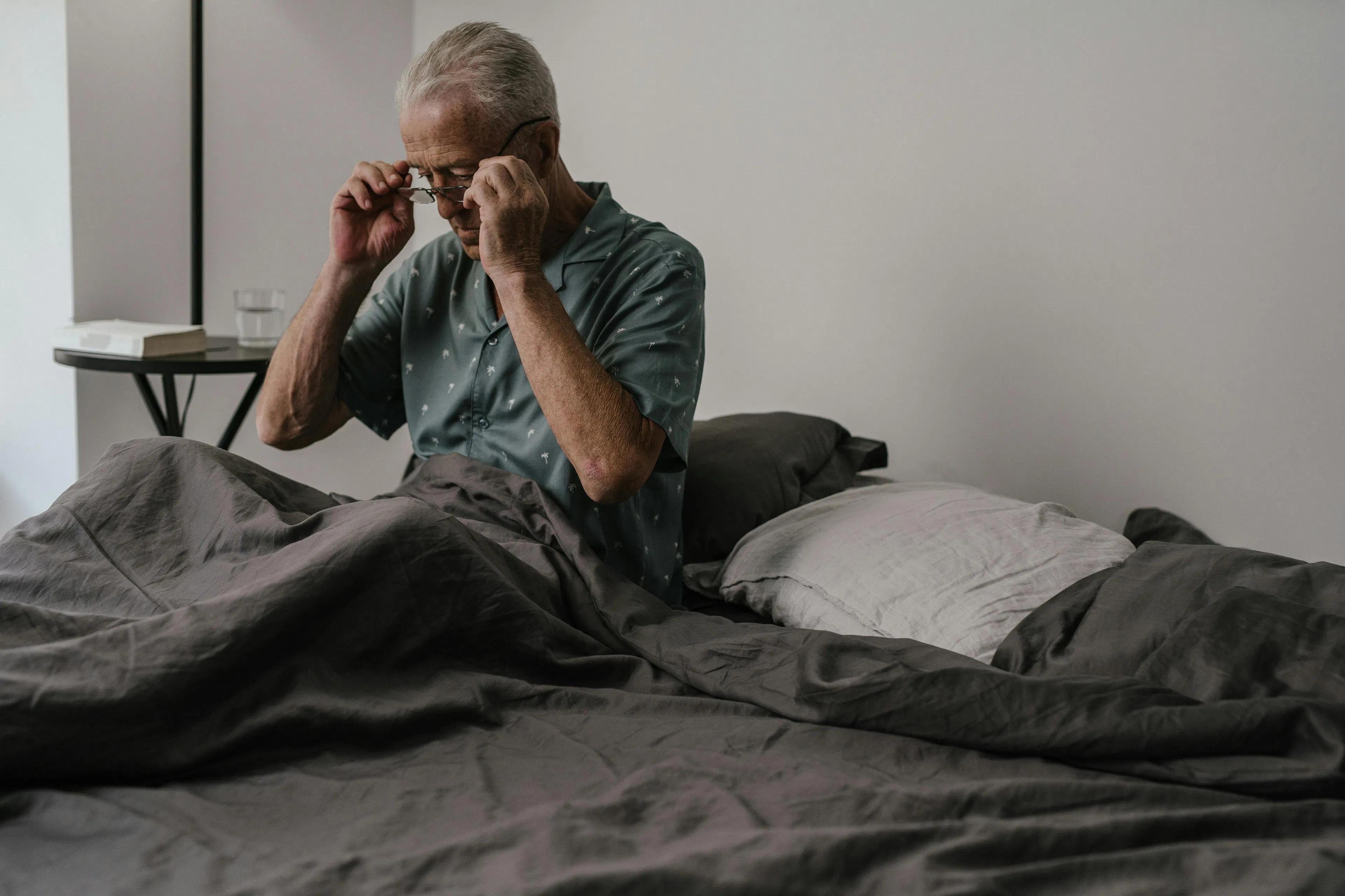 A man sitting on the edge of a bed to wipe sweat from his forehead