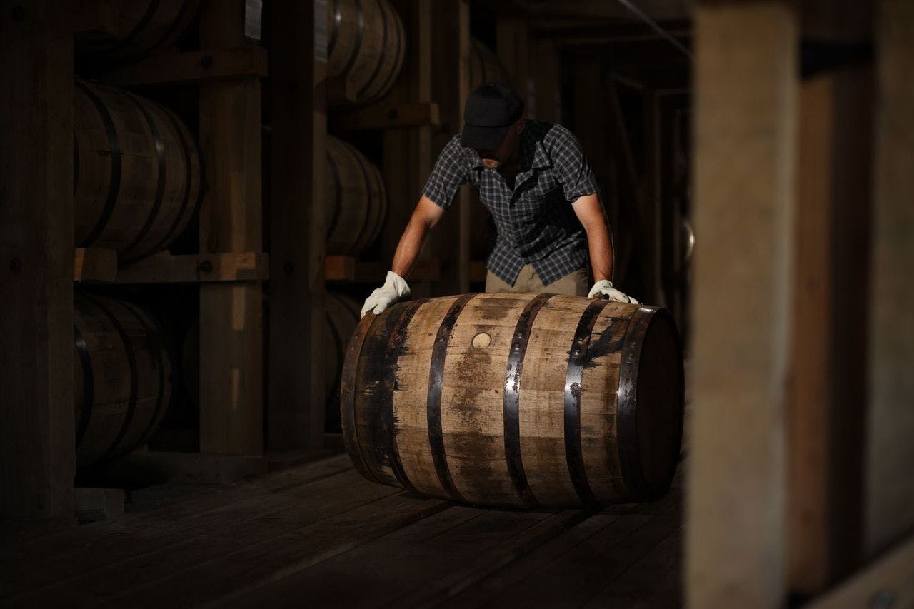A man rolling a wooden barrel through a racked bourbon warehouse.