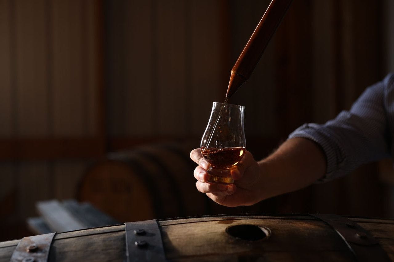 A man pouring Kentucky bourbon into a glass, with a barrel visible in the background.