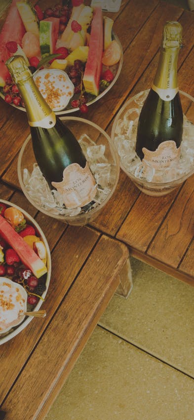 Champagne bottles in buckets filled with ice, alongside bowls of mixed fruits on a table.