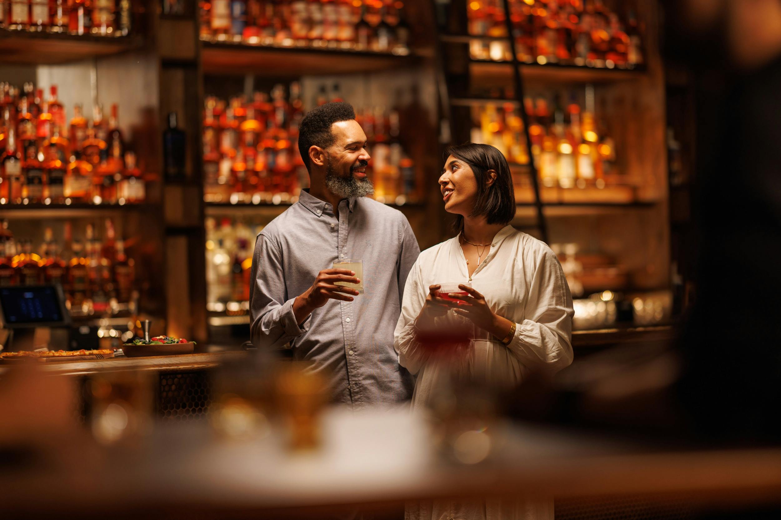 Two people enjoying their bourbon, with a display of liquor bottles behind them.