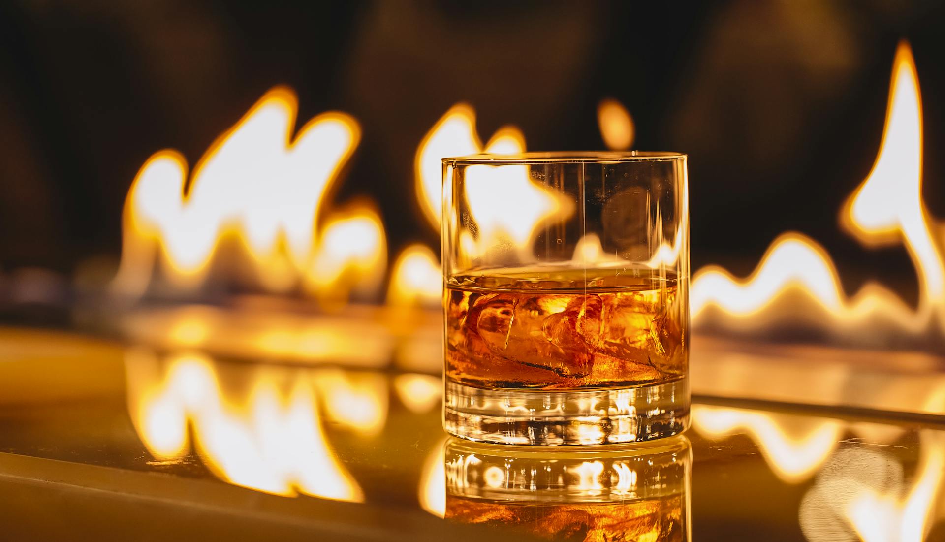 A glass of bourbon in sharp focus in the foreground, with a dramatic low flame flickering in the blurred background, creating a warm and moody ambiance.