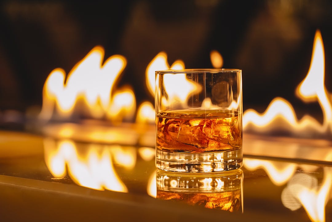 A glass of bourbon in sharp focus in the foreground, with a dramatic low flame flickering in the blurred background, creating a warm and moody ambiance.