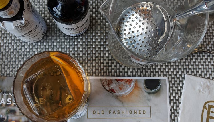A table with bar essentials: a glass of whiskey on the rocks, a couple of bottled liquors, and some bar tools.