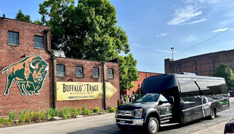 A Mint Julep Experiences luxury vehicle parked in front of Buffalo Trace Distillery, with the historic brick buildings and iconic signage visible in the background.