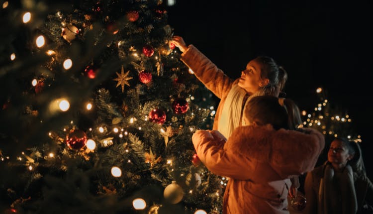 A woman and a child decorating a Christmas tree, while another child watches from the background.
