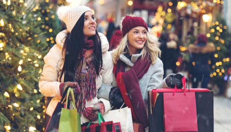 Two women in winter outfits carrying colorful shopping bags, set against a festive Christmas background.
