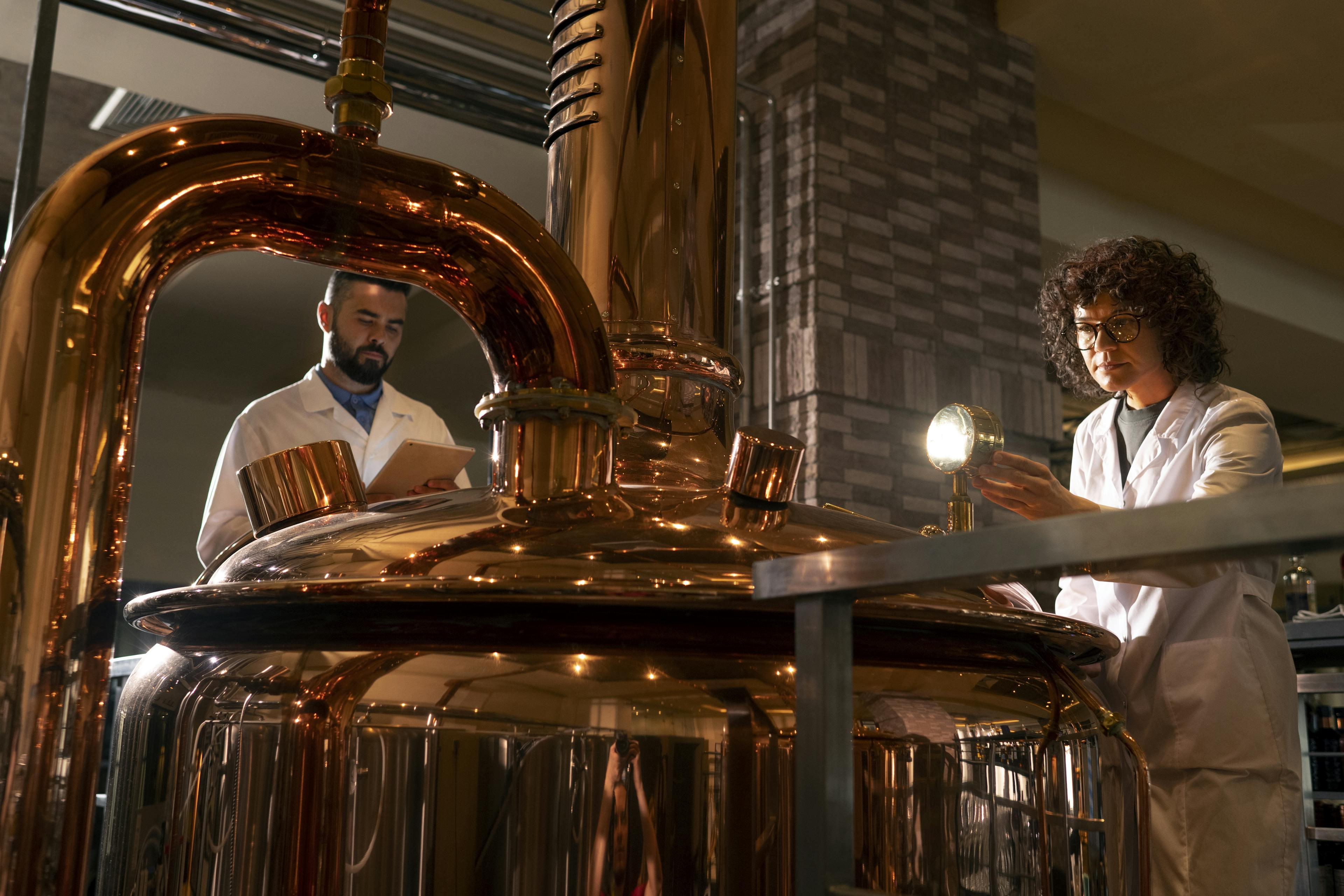 Two distillery workers standing around a copper still, inspecting it carefully.