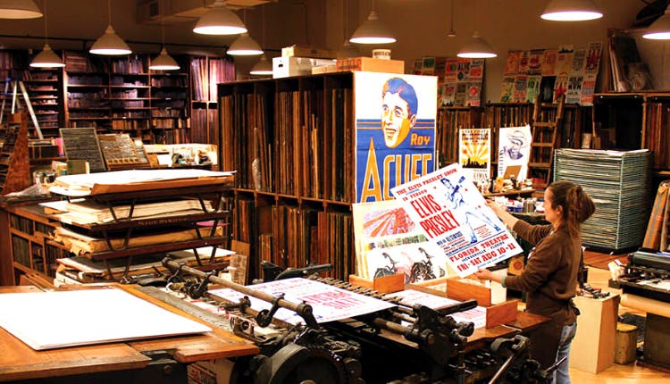 The interior of Hatch Show Print in Nashville, showing a woman holding a print, would feature the vibrant and historic atmosphere of the print shop