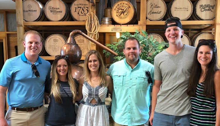 A group of tourists smiling and posing for a photo, with a wall of stacked wooden whiskey barrels behind them, showcasing the rustic charm of a distillery setting.A group of tourists smiling and posing for a photo, with a wall of stacked wooden whiskey barrels behind them, showcasing the rustic charm of a distillery setting.