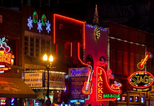 Broadway Street in downtown Nashville at night, illuminated by the vibrant glow of neon lights from various bars, restaurants, and live music venues.
