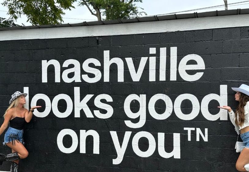 Two women happily posing in front of a "Nashville Looks Good on You" mural.
