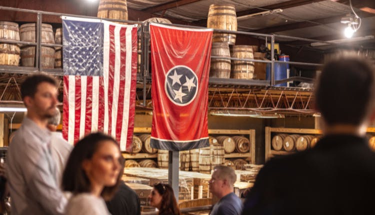 The interior of Pennington Distilling Co.'s rickhouse, with tourists exploring, while focusing on the USA and Tennessee flags displayed prominently.