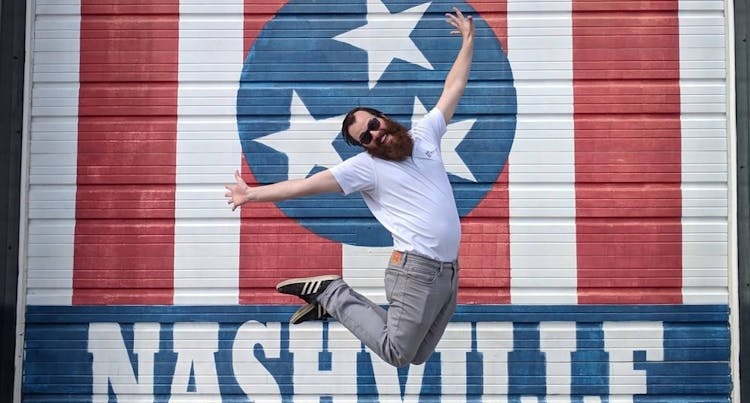 A man joyfully jumping in front of a Nashville mural featuring red and white stripes with stars.