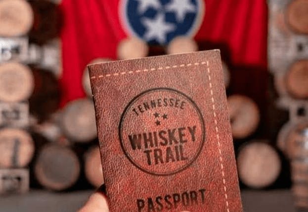 A Tennessee Whiskey Trail Passport held in focus, with a blurred background of stacked wooden whiskey barrels.
