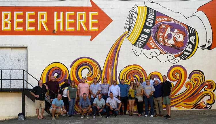 A group of tourists posing in front of a large wall mural that says "Beer Here," featuring an artsy illustration of beer pouring from a can.