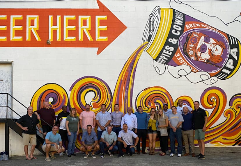 A group of tourists posing in front of a large wall mural that says "Beer Here," featuring an artsy illustration of beer pouring from a can.