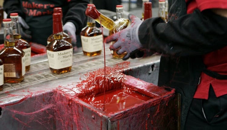 A gloved hand holding a Maker’s Mark bottle, preparing it for waxing during an interactive distillery tour experience.