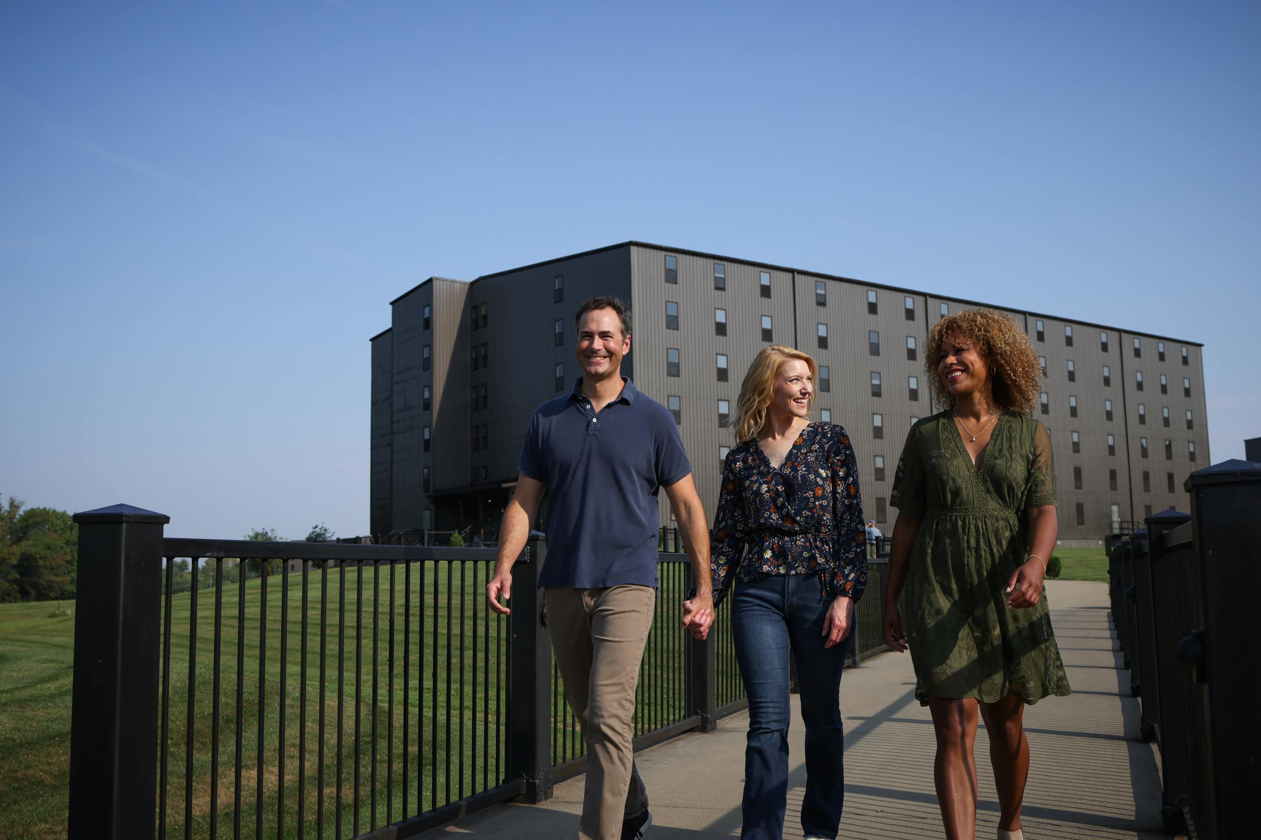 Two women and a man walk the grounds of a distillery with a bourbon barrel aging house in the background.