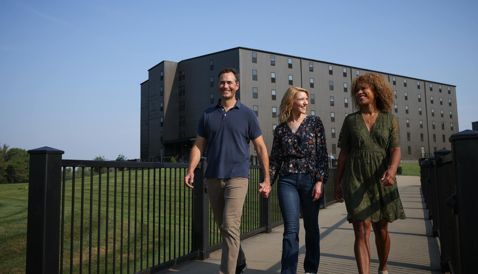 Two women and a man walk the grounds of a distillery with a bourbon barrel aging house in the background.
