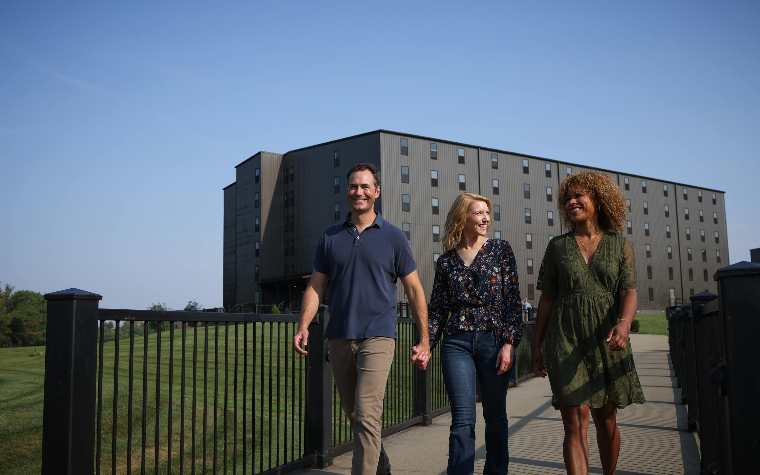 Two women and a man walk the grounds of a distillery with a bourbon barrel aging house in the background.
