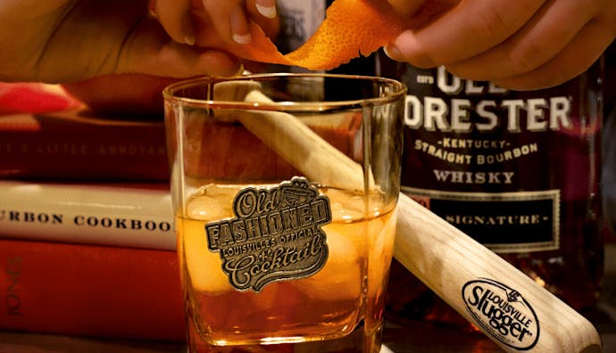A glass of Old Forester Whiskey, with hands slightly out of frame holding a twist of citrus peel.