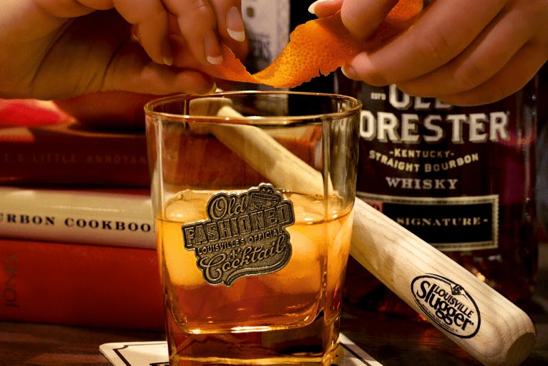 A glass of Old Forester Whiskey, with hands slightly out of frame holding a twist of citrus peel.