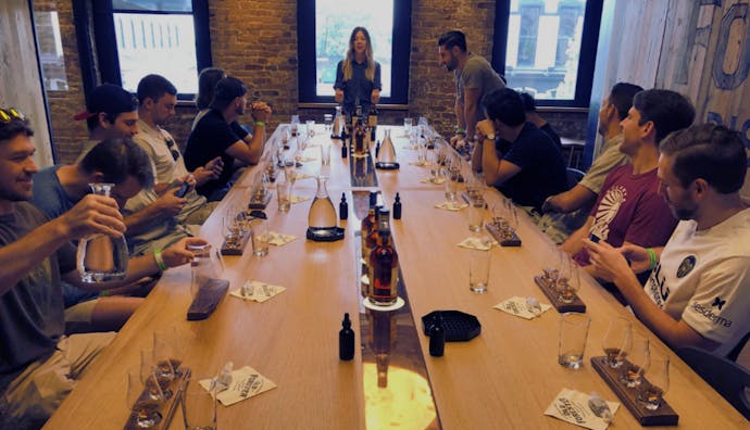 A group of tourists sits at a table, each with a set of glasses containing whiskey samples. A facilitator stands in front, speaking to the group.