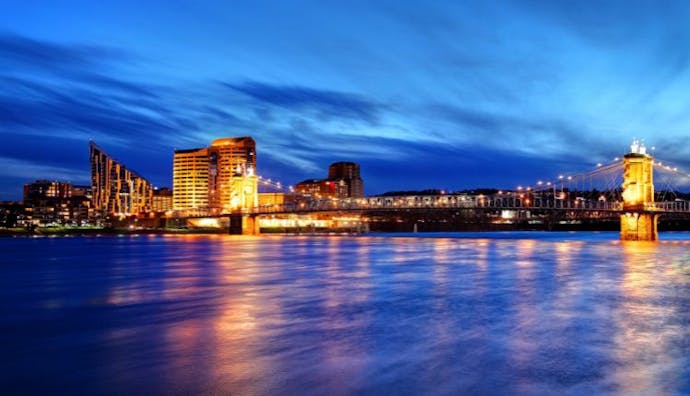 Covington, Kentucky at night, featuring the iconic John A. Roebling Suspension Bridge and surrounding buildings brightly lit with amber lights, set against a dark blue sky and dark blue water in the foreground.