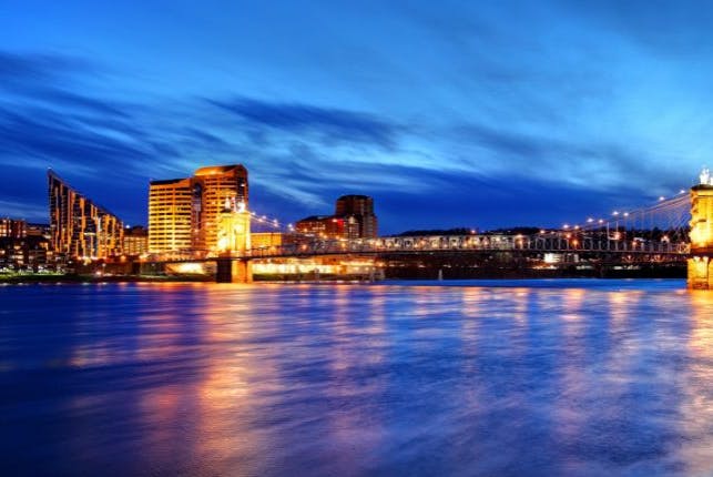 Covington, Kentucky at night, featuring the iconic John A. Roebling Suspension Bridge and surrounding buildings brightly lit with amber lights, set against a dark blue sky and dark blue water in the foreground.