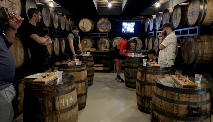 A group of people are standing beside individual whiskey barrels in a rickhouse.