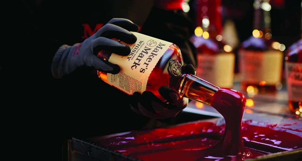 A gloved hand dipping a Maker's Mark bottle into red wax.