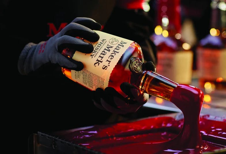 A gloved hand dipping a Maker's Mark bottle into red wax.