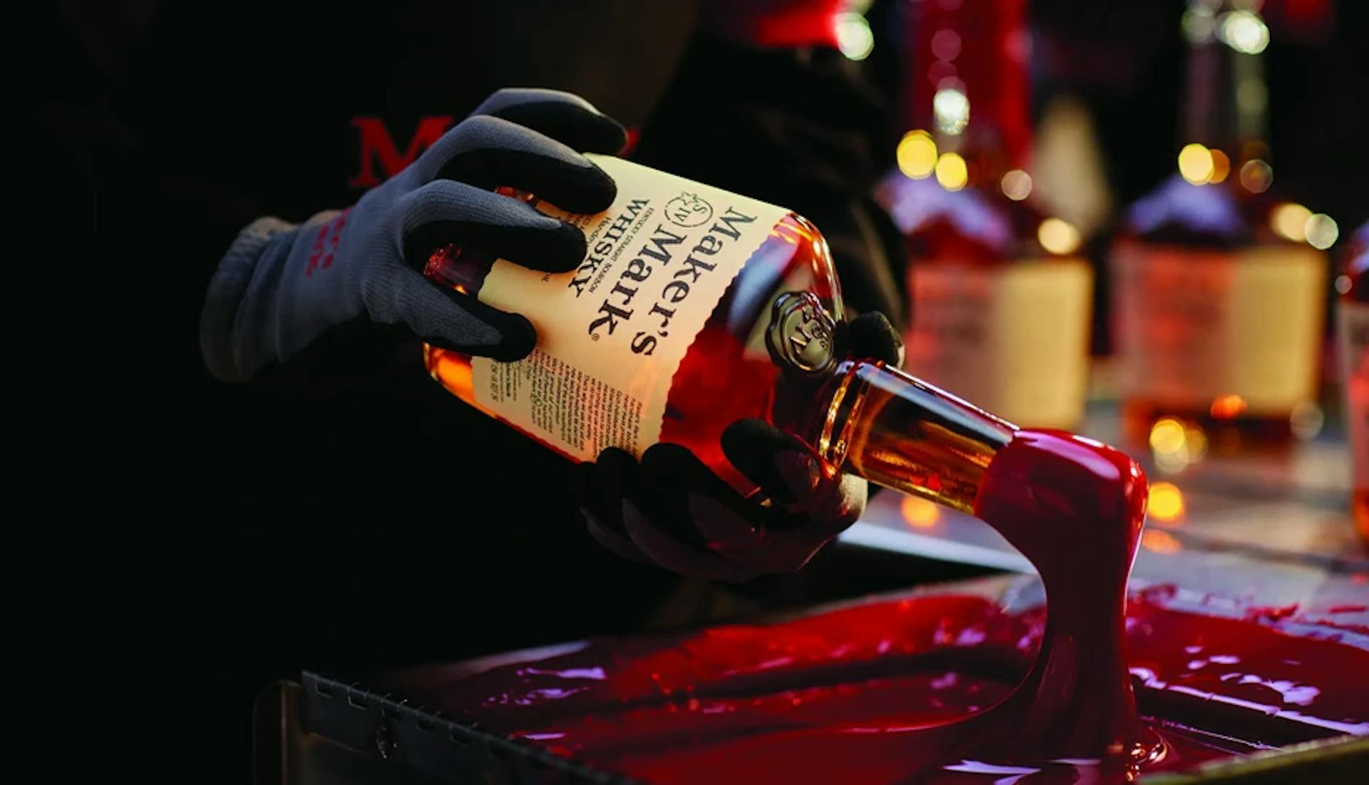 A gloved hand dipping a Maker's Mark bottle into red wax.